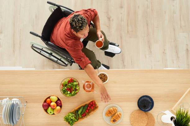 High angle view of disabled woman in wheelchair holding cup of tea in her hand and reaching her hand for the buns on the table